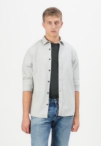 Camisa de botón gris claro, con cuello, mangas remangadas y botones negros, llevada sobre una camiseta negra ajustada y jeans azules.