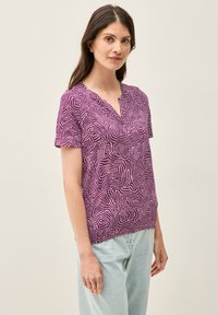 Top de manga corta, de color púrpura, con un patrón blanco texturizado y ondulado. Cuenta con un escote en V y un corte relajado, fabricado en material ligero.