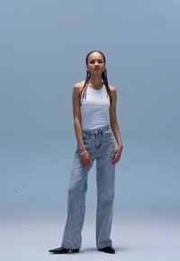 Top branco de alças e jeans de lavagem clara, ajustados na cintura, com a parte inferior a abrir, usados com sapatos pretos de bico fino. Design simples e elegante.