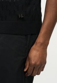 Χέρι με σκούρο δέρμα ακουμπά κοντά στην τσέπη μαύρου παντελονιού, φορώντας μαύρο πλεκτό τοπ με υφή και μικρό καρφίτσα με το λογότυπο "KL".