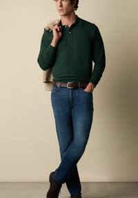 Tmavě zelená dlouhá polokošile v kombinaci s modrými džíny. Hnědý kožený opasek a boty. Světlezelený svetr přehozený přes jedno rameno.