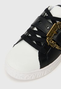 Biele a čierne tenisky s čiernymi šnúrkami, dekoratívnym zlatým reťazcom a nápisom "Versace Jeans Couture" vytlačeným na bielej podrážke.