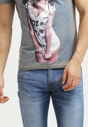 Mann trägt blaue Jeans und ein graues T-Shirt mit einem aufgedruckten Bild einer Frau in weißem Outfit auf der Vorderseite.