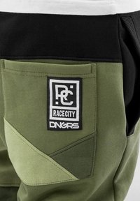 Pantaloni de trening verde măsliniu cu un brâu negru, accente de design triangular și o etichetă cu patch cu brandul "Race City" și "DNGRS".