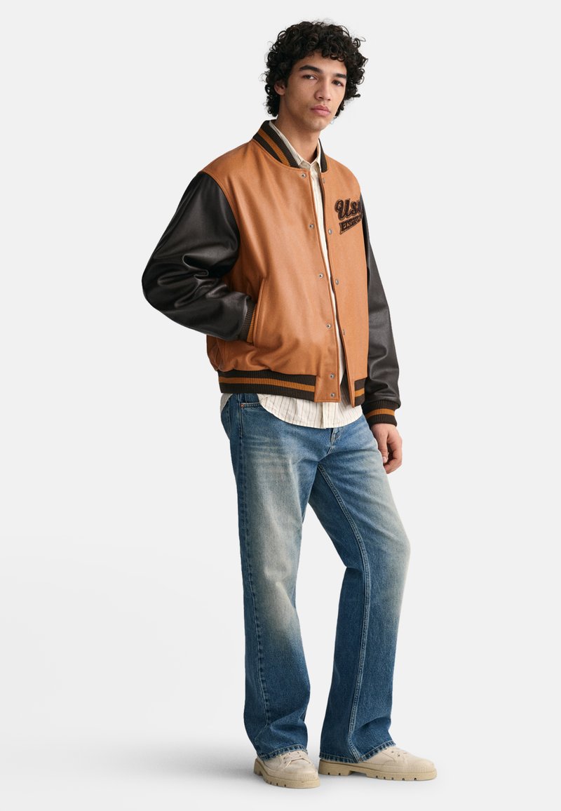 Braune und schwarze Collegejacke mit Druckknöpfen, einem gerippten Kragen und einem Logo auf der Brust, kombiniert mit hellblauen Jeans und beigen Schuhen.