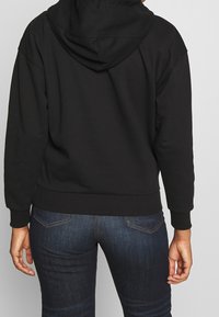 Černá mikina s kapucí na šňůrku, žebrovanými manžetami a lemem a hladkým povrchem. Doplněná tmavě modrými džíny.