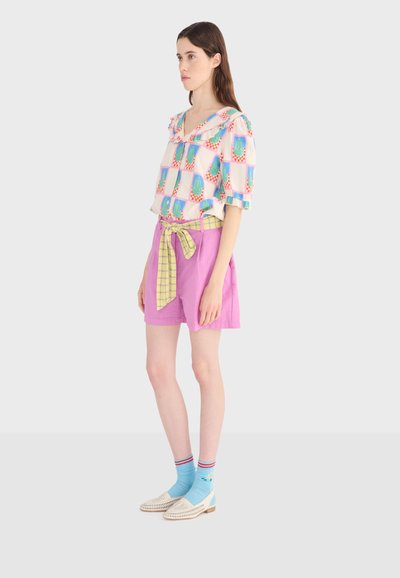 Jeune femme portant un chemisier pastel à motifs, un short rose avec une ceinture à carreaux jaune, des chaussettes bleues et des chaussures blanches à enfiler, debout de profil.
