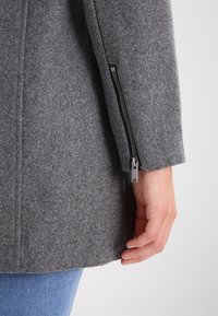 Šedý vlněný kabát se zipem na manžetě, ukazující texturovanou látku a čisté linie, částečně nošený přes modrý denim.