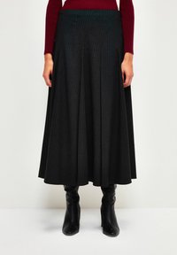 Jupe noire plissée longue jusqu'au sol avec un subtil motif à rayures verticales, portée avec des bottes noires ajustées jusqu'aux genoux et un haut bordeaux.