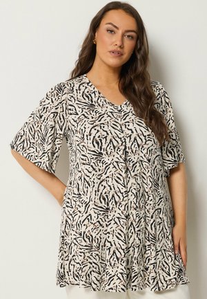 Kvinde med langt bølget hår iført en løs, kortærmet, beige kjole med sort abstrakt dyreprint, poserer med den ene hånd på hoften.
