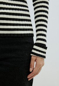 Schwarz-weiß gestreiftes, geripptes Langarmshirt mit umgeschlagenen Manschetten, kombiniert mit dunkler Jeans. Nahaufnahme des Ärmels und der Hand.