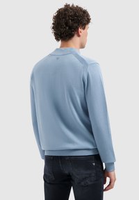 Man met krullend haar, gekleed in een lichtblauwe langekleurige trui en donkere jeans, met zijn rug naar de camera toe tegen een effen lichtgrijze achtergrond.