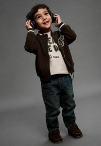 Jongen in een donkerbruin zip-up jacket, witte graphic t-shirt, blauwe jeans en bruine schoenen, met een hoofdtelefoon in zijn hand, glimlachend tegen een grijze achtergrond.