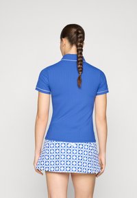 Polo shirt bleu à côtes avec des accents blancs, associé à une jupe blanche à motif circulaire bleu et jaune. Vue de dos présentée.