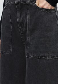 Personne portant un jean noir ample avec de grandes poches avant plaquées, main partiellement glissée dans la poche droite, haut noir visible.