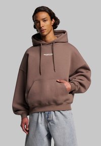 Braune übergroße Kapuzenjacke mit frontaler Tasche und Kordelzugkapuze. Zeigt dezente Markenkennzeichnung in Weiß auf der Brust. Kombiniert mit hellblauen Jeansshorts.