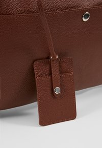 Sac en cuir marron texturé avec une étiquette rectangulaire en cuir attachée, sécurisée par un bouton-pression argenté sur fond blanc.