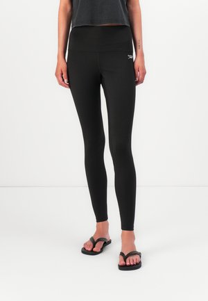 Zwarte leggings gemaakt van rekbaar materiaal, met een hoge tailleband. Gecombineerd met slippers en een kort, donker topje. Eenvoudig ontwerp.
