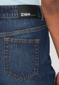 Egy ember csípőjén látható kék farmernadrág hátsó zsebe, amelyen jól látható narancssárga varrás és fekete címke olvasható: "ZIGN STUDIO".
