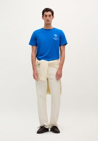 Blå bomuld t-shirt med hvidt logo, cremefarvede bukser og brune sko. T-shirten har en rund hals; bukserne har en afslappet pasform med snoretræk.