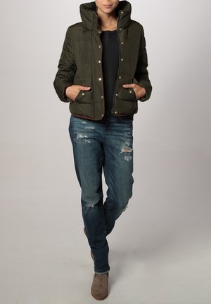 Femme portant une veste matelassée vert foncé, un haut noir, un jean bleu déchiré et des bottines en daim marron, debout les mains dans les poches.