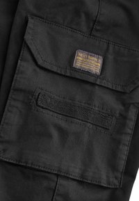 Primer plano del bolsillo de tela negra de unos pantalones con solapa, tira de velcro y pequeña etiqueta rectangular con el texto "Next Supply" en color amarillo.