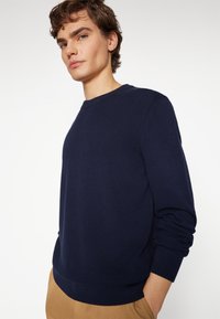 Maglione lavorato a maglia blu navy con scollo rotondo e polsini a costine. Presenta un tessuto testurizzato e un design slim fit. Abbinato a pantaloni beige.