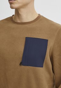 Sudadera de felpa marrón con un bolsillo parche azul marino y detalle de cremallera. Cuello redondo y tejido texturizado para mayor calidez.