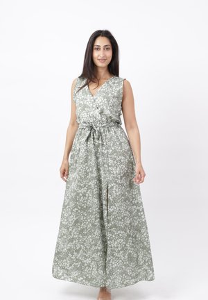 Kvinde stående iført ærmeløs grå maxikjole med hvidt blomsterprint og taljebånd, smilende mod hvid baggrund.