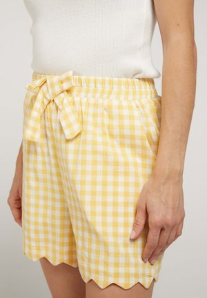 Shorts à carreaux vichy jaunes et blancs avec ourlet festonné et nœud à la taille, portés avec un haut blanc sans manches.