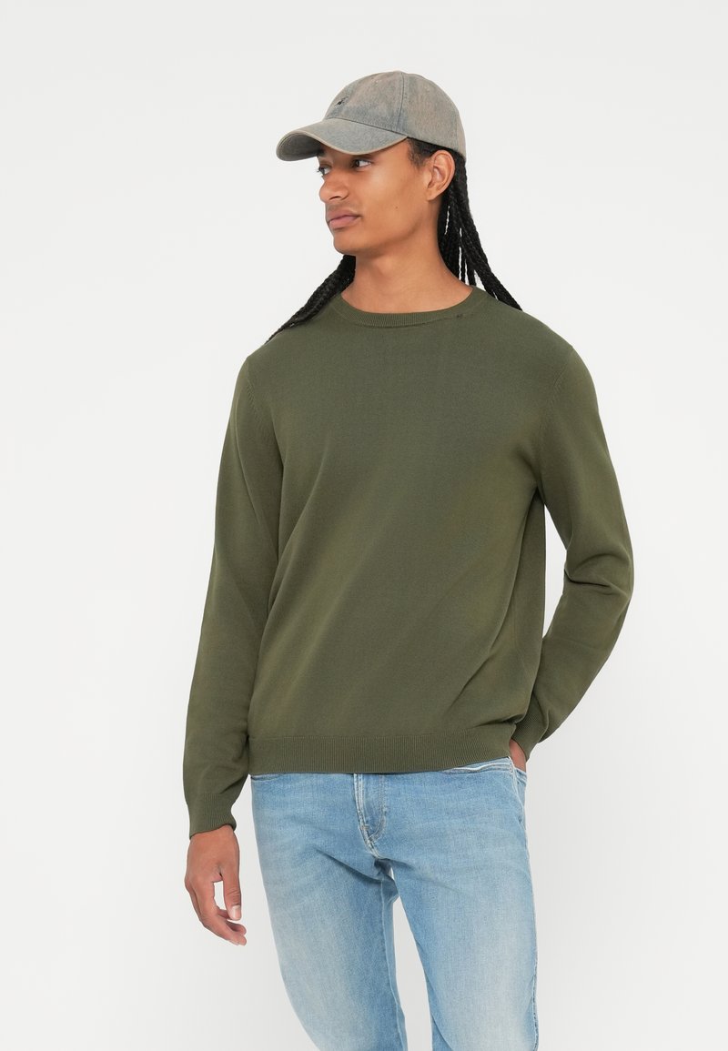 Olivgrüner Strickpullover mit Rundhalsausschnitt, kombiniert mit hellblauen Jeans und einer grauen Baseballkappe. Der Stoff wirkt glatt und körpernah.