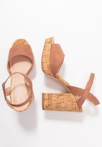 Hnědé suédé platformové sandály s korkovými podpatky. Mají popruhy kolem kotníku a křížový design. Hladký povrch s přírodním korkovým vzhledem.