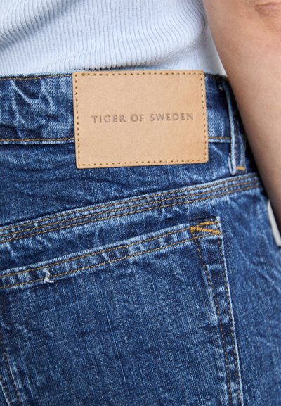 Tumši zili džinsi ar teksturētu apdari, ar brūnu ādas uzšuvi, kurā rakstīts "TIGER OF SWEDEN". Džinsu kabatām kontrastējošs šuvums.