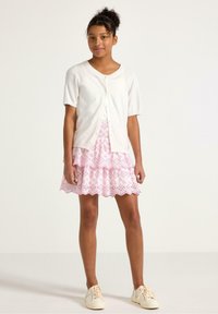 Cardigan bianco a maniche corte con chiusura a bottoni, abbinato a una gonna a più strati con motivo floreale rosa. Sneakers leggere completano il look.