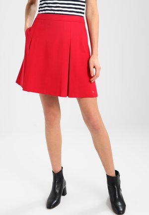 Jupe plissée rouge en tissu lisse, avec poches latérales et taille élastiquée. Associée à des bottines noires pour un contraste.