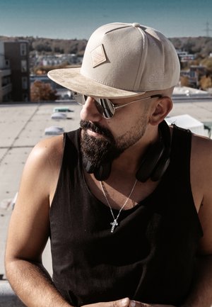 Beige baseball cap with a textured finish, silver aviator sunglasses, black tank top, and black headphones. Simple silver cross necklace.