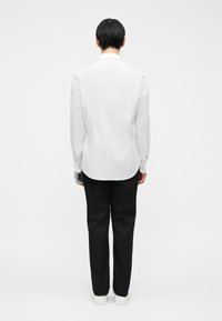 Άτομο που στέκεται με την πλάτη στραμμένη, φορώντας λευκό μακρυμάνικο πουκάμισο, μαύρο παντελόνι και λευκά αθλητικά παπούτσια σε απλό λευκό φόντο.