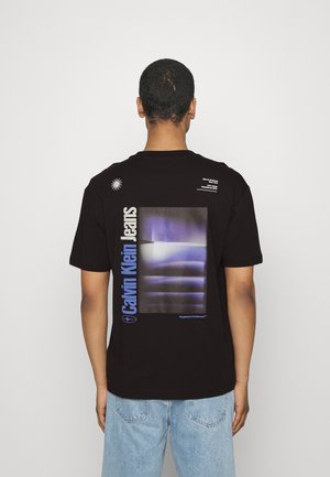 Mann trägt ein schwarzes Calvin Klein Jeans T-Shirt mit blauem und weißem Grafik- und Textmotiv auf der Rückseite, kombiniert mit hellblauen Jeans vor einfärbigem Hintergrund.