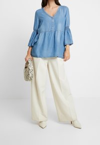 Light blue denim top with a V-neck and three-quarter bell sleeves, paired with cream wide-leg trousers and a patterned handbag.