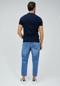 Camisa de manga corta azul marino, jeans de color azul claro y zapatillas blancas con acentos en azul marino, vistas desde la parte posterior sobre un fondo neutro.