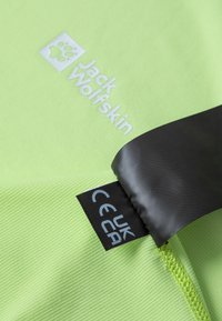 Hellgrüner Stoff mit strukturiertem Oberflächen verfügt über ein weißes "Jack Wolfskin"-Logo. Ein schwarzes Etikett enthält das CE-Zeichen und Pflegehinweise.