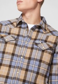 Camicia in flanella a quadri marrone, blu e beige con due tasche sul petto, bottoni a pressione e un'etichetta con logo Wrangler sulla tasca sinistra.