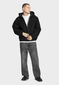 Czarna casualowa kurtka z kapturem, elastycznymi mankietami i zapięciem na zamek, nałożona na białą koszulkę i w zestawie ze spodniami dżinsowymi w odcieniu wyblakłego szarego.