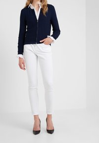 Cárdigan azul marino de canalé con cremallera y mangas largas, combinado con pantalones blancos ajustados y tacones altos negros, estilizado con una camisa blanca con cuello debajo.