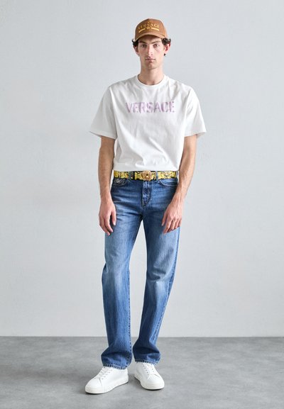 Weißes T-Shirt mit "VERSACE"-Schriftzug, blaue Jeans, goldgemusterter Gürtel, beiger Hut mit Logo, weiße Sneakers, stehend vor einem grauen Hintergrund.
