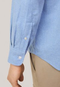 Camisa de manga larga azul claro hecha de una tela con sutil textura, que presenta puños abotonados con botones en contraste y un dobladillo curvado.
