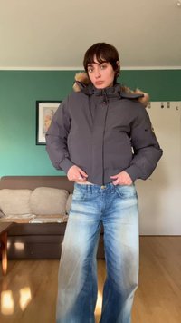 Casaco puffer cinza com capuz forrado a pelo, apresentando um fecho frontal e punhos canelados. Combinado com jeans de perna larga em azul desbotado.
