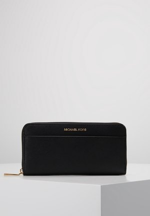 Portefeuille noir à fermeture éclair avec tirette en métal doré et logo "Michael Kors" embossé en doré sur le devant, posé sur une surface blanche avec un fond uni.