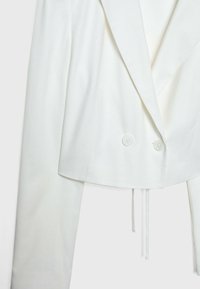 Blazer blanco recortado con solapa, cierre de doble botón y detalle de lazo en la cintura; hecho de una tela suave y con textura.
