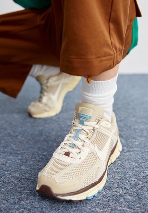 Beżowe buty sportowe Nike Vomero 5 noszone z białymi skarpetkami i brązowymi spodniami, stojące na teksturowanym niebieskim dywanie.